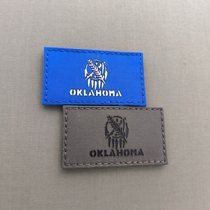 May include: Two embroidered patches with the state seal of Oklahoma. The top patch is blue with white stitching and the word "OKLAHOMA" in white. The bottom patch is brown with black stitching and the word "OKLAHOMA" in black.