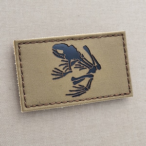 Pode incluir: Um remendo marrom com um esqueleto de sapo bordado em preto. O remendo tem costuras marrons ao redor das bordas.