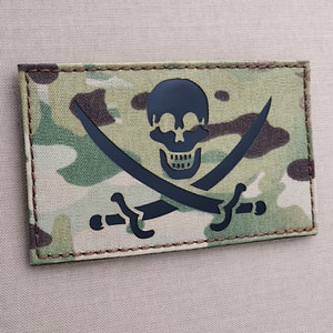 May include: A black and white embroidered patch with a skull and crossbones design on a camouflage background. The patch is sewn onto a beige fabric.