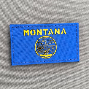 May include: A blue rectangular patch with the word "MONTANA" in yellow block letters. Below the text is a yellow circular emblem with a landscape design. The patch is stitched with black thread.