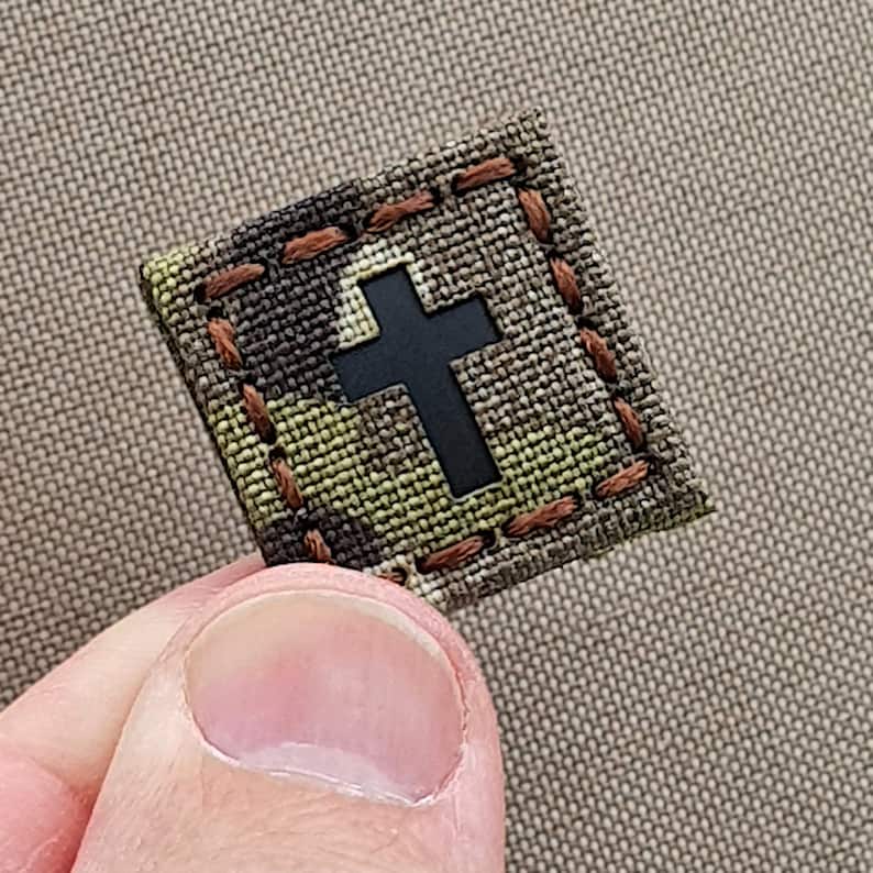 K&ouml;nnte beinhalten: Ein kleiner, quadratischer Patch mit einem Tarnmuster und braunen N&auml;hten. Ein schwarzes Kreuz ist mittig auf dem Patch platziert.