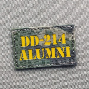 Puede incluir: Un parche de camuflaje verde, marrón y negro con costuras amarillas. El parche tiene el texto "DD-214 ALUMNI" en amarillo.
