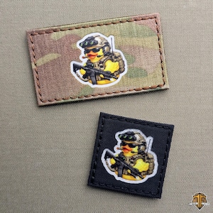 Op de afbeelding: Twee tactische patches. Eén heeft een camouflage achtergrond, de andere is zwart. Elk toont een gele eend in militaire uitrusting, met een geweer en een nachtzichtbril en een helm.