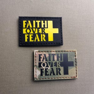 Könnte beinhalten: Zwei rechteckige Aufnäher mit dem Text "FAITH OVER FEAR". Ein Aufnäher ist schwarz mit gelbem Text und einem gelben Kreuz. Der andere Aufnäher hat ein Tarnmuster mit schwarzem Text und einem schwarzen Kreuz. Beide Aufnäher befinden sich auf einem beigen Hintergrund.