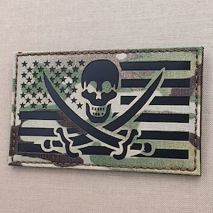 May include: Black and white embroidered patch with a skull and crossbones design on a camouflage background. The skull and crossbones are superimposed on a stylized American flag.