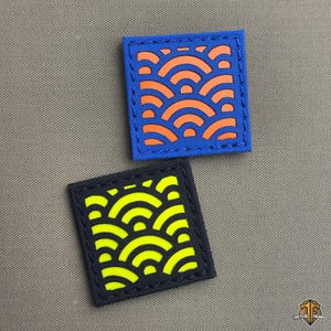 Op de afbeelding: Twee vierkante patches met een golfpatroon. Eén patch is blauw met oranje golven, de andere zwart met gele golven. Beide patches hebben een gestikte rand. De achtergrond is grijs.