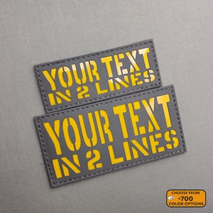 Può includere: Due toppe rettangolari grigie con testo giallo che recita "YOUR TEXT IN 2 LINES". Le toppe sono cucite lungo i bordi. Un piccolo rettangolo arancione in basso a destra dice "CHOOSE FROM +700 COLOR OPTIONS".