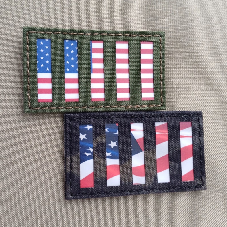 K&ouml;nnte beinhalten: Zwei amerikanische Flaggen-Patches, einer mit gr&uuml;nem Hintergrund und der andere mit schwarzem Tarnmuster-Hintergrund. Beide Patches haben sechs vertikale Streifen mit einem gen&auml;hten Rand.