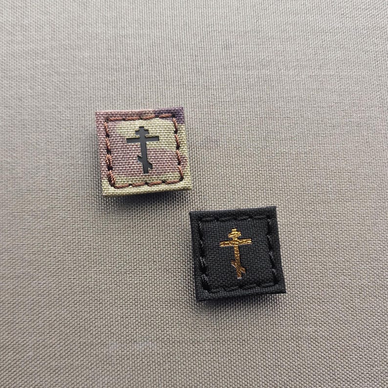 K&ouml;nnte beinhalten: Zwei kleine, quadratische Stoffaufn&auml;her mit gestickten Kreuzen. Einer hat ein Camouflagemuster mit einem schwarzen Kreuz, der andere ist schwarz mit einem goldenen Kreuz. Beide haben gen&auml;hte Kanten.