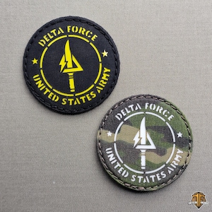 Könnte beinhalten: Zwei runde Aufnäher mit den Aufschriften "DELTA FORCE" und "UNITED STATES ARMY". Einer ist schwarz mit gelbem Text und einem gelben Pfeilsymbol. Der andere hat ein Tarnmuster mit weißem Text und einem weißen Pfeilsymbol.