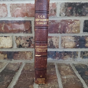 May include: A vintage, brown leather-bound book from the I.C.S. Reference Library. The spine features gold lettering with the title and the words "Arithmetic Elementary Drawing". The book is set against a brick background.