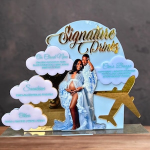 May include: A blue and gold acrylic sign with the text "Signature Drinks" in gold lettering. The sign features a cloud with the text "On Cloud Nine" and a list of ingredients, a cloud with the text "Sunshine" and the text "Non-Alcoholic Punch", a cloud with the text "Other" and the text "Soda, Water, Wine, Beer", and a plane with a cloud with the text "Crun's Booze" and a list of ingredients.