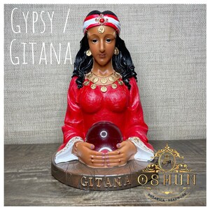 Op de afbeelding: Een rood en gouden beeldje van een vrouw met een rode tulband en een kristallen bol in haar handen. Het beeldje is gelabeld "Gitana Oshun Botanica Beadwork".