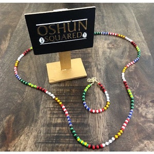 Peut inclure: Un ensemble de collier et de bracelet en perles colorées avec une pancarte indiquant "OSHUN SQUARED".