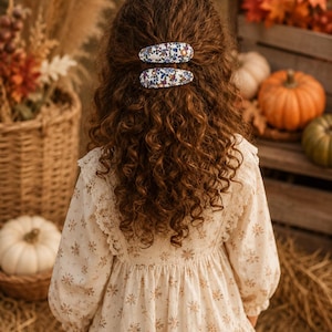 May include: Two oval hair clips featuring a dense floral pattern with blue, red, and green blossoms on a white background. The clips are positioned one above the other, securing a section of long, wavy, dark brown hair at the back of the head.
