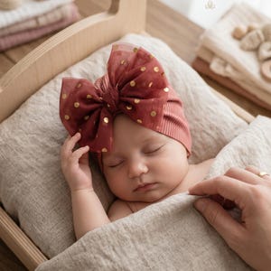 Könnte beinhalten: Ein gerippter Babyturban in Altrosa mit einer großen, transparenten, dunkelroten Tüllschleife, die mit metallisch goldenen Punkten verziert ist. Der Text LITTLE SPLENDOR - Elegant Comfort for your Everyday - ist oben rechts zu sehen.