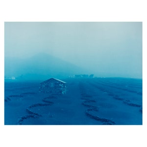 May include: A single-story stone building with a roof stands in a field of blue grass. The building is surrounded by a series of winding paths. The background is a hazy blue sky.