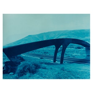 May include: A cyanotype print of a modern bridge spanning a highway. The bridge has a curved, dark blue arch supported by two pillars. The landscape features a mountain range and sparse vegetation, all in shades of blue.