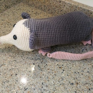 May include: A handmade crochet opossum plush toy. The opossum has a gray body, a white face, and a pink nose and tail. The toy has black button eyes and small gray ears. The plush is approximately 12 inches long.