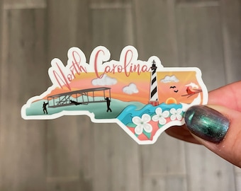 May include: A sticker shaped like the state of North Carolina with a sunset, a lighthouse, a plane, and flowers. The text "North Carolina" is written in pink cursive.