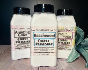 May include: Three white plastic bottles with black lids and labels. The labels are for carpet freshener with different scents: Amazing Grace, Beachwood, and Lemon & Lavender. The bottles are on a wooden surface with a green and white flower in the background.