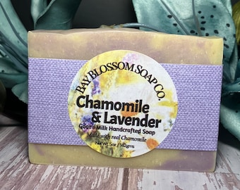 May include: A bar of goat's milk soap with a lavender and chamomile scent. The soap is wrapped in a purple label with the text "BAY BLOSSOM SOAP CO. Chamomile & Lavender Goat's Milk Handcrafted Soap Made with real Chamomile Net Wt. 5oz / 142gms".