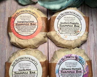 May include: Four round bars of soap wrapped in brown paper. Each bar has a white label with the text "Bay Blossom Soap Co." and the name of the scent: "Energizing Citrus", "Southern Magnolia", "Vanilla Mint", and "Saltwater Mermaid". Each bar is labeled as a "Shampoo Bar" and is pH balanced and soap free.