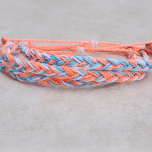 Puede incluir: Pulseras trenzadas en tonos melocotón, azul claro y blanco. Las pulseras están hechas de cordón tejido, con una trenza central más gruesa y cordones más finos a juego. Las pulseras se exhiben sobre una superficie neutra.