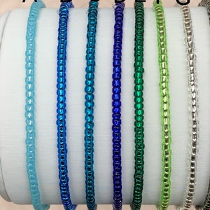 May include: Seven beaded bracelets in various colors, including light blue, blue, teal, dark blue, green, light green, and silver. The bracelets are arranged in a row on a white surface.