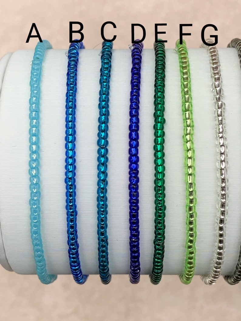 May include: Seven beaded bracelets in various colors, including light blue, blue, teal, dark blue, green, light green, and silver. The bracelets are arranged in a row on a white surface.
