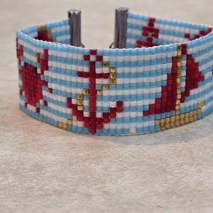 May include: A wide beaded cuff bracelet featuring a nautical theme with horizontal light blue and white stripes. The design includes red crab, anchor, and sailboat motifs accented with small metallic gold beads and silver-toned metal end bars.