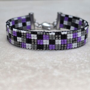May include: A woven bead bracelet featuring a repeating checkered pattern in shades of purple, black, white, and gray. The bracelet is finished with silver-toned metal end caps and a lobster claw clasp, creating a structured and uniform design.