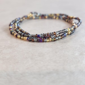May include: A multi-strand beaded bracelet with a mix of metallic and iridescent beads in shades of gold, silver, brown, purple, and blue. The bracelet is coiled on a neutral surface, showcasing its intricate design.