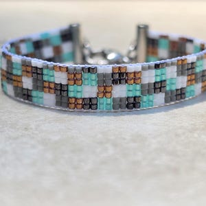 May include: A woven bead bracelet featuring a repeating checkered pattern in alternating colors of white, turquoise, metallic copper, and dark charcoal gray. The bracelet is finished with silver-toned metal end caps and a lobster claw clasp.