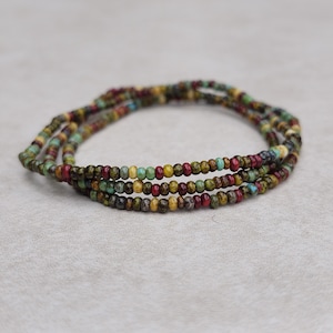May include: Three strands of beaded bracelets in earthy tones. The beads are small and round, with colors including red, green, yellow, and blue. The bracelets are arranged in a circular shape, resting on a light gray surface.