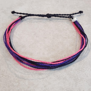 May include: Adjustable bracelet featuring a combination of pink, purple, black, and white threads. The bracelet has a black adjustable cord and a small silver-colored charm. The threads are tightly woven together to form the bracelet.