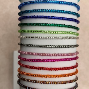 May include: A stack of 14 different colored beaded bracelets. The bracelets are all made with small, round beads and are arranged in a vertical stack. The colors of the bracelets include light blue, blue, dark blue, green, light green, silver, gray, pink, red, orange, gold, brown, and purple.