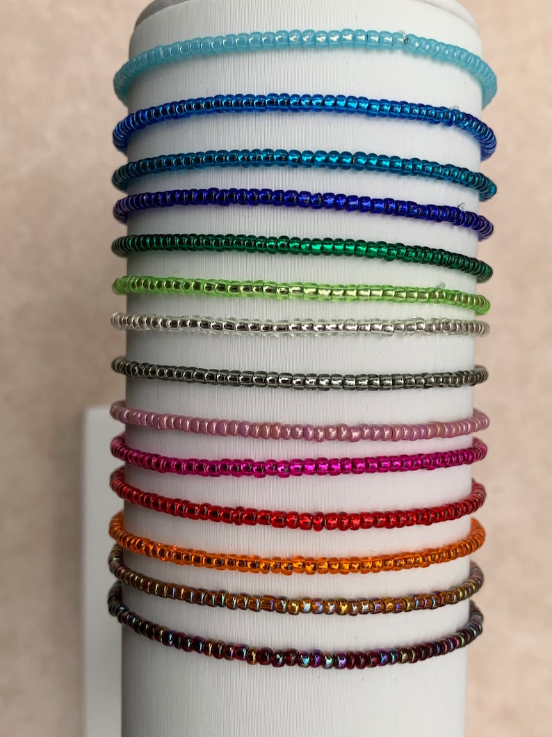 May include: A stack of 14 different colored beaded bracelets. The bracelets are all made with small, round beads and are arranged in a vertical stack. The colors of the bracelets include light blue, blue, dark blue, green, light green, silver, gray, pink, red, orange, gold, brown, and purple.