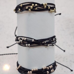May include: Three black string bracelets with metallic beads. The top bracelet has gold beads, the middle one has silver beads, and the bottom one has rose gold beads. Each bracelet is wrapped around a white cylinder.