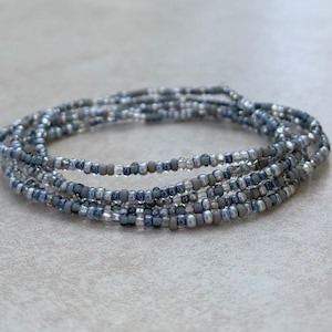 May include: A stack of beaded bracelets in shades of gray, silver, and clear. The bracelets are made of small, round beads strung together, creating a delicate and layered look. The beads have a subtle sheen.