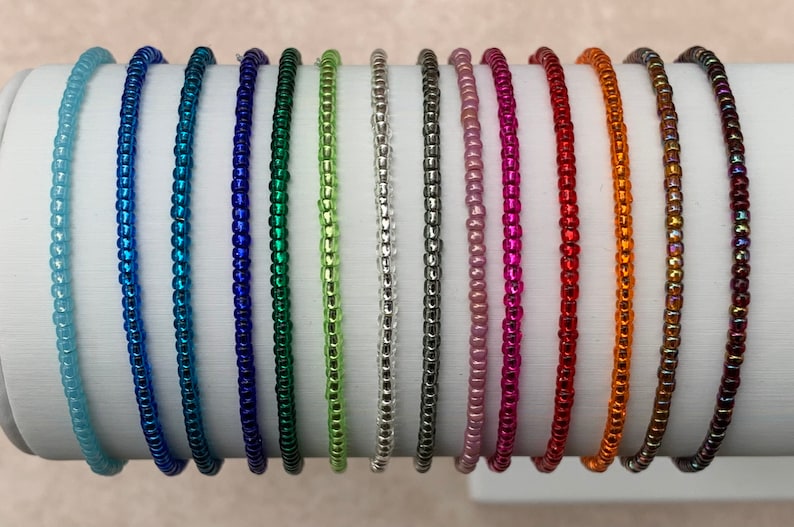 May include: A collection of 14 colorful beaded bracelets, each with a different color and finish. The bracelets are arranged in a row on a white surface.