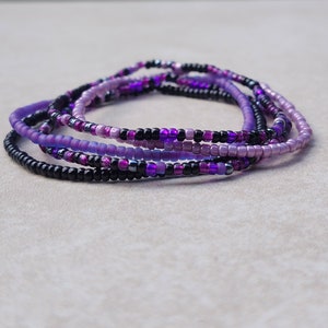 May include: A set of five beaded bracelets in shades of purple and black. The bracelets are made with small, round beads and are stacked on top of each other.