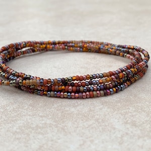 May include: A multi-strand beaded bracelet in shades of brown, orange, and purple. The tiny beads are strung together to create a flexible, wrap-around design. The bracelet is shown coiled on a neutral surface.