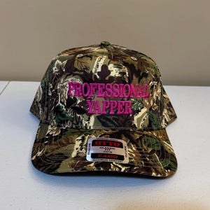 May include: Camouflage baseball cap with the words "Professional Yapper" embroidered in pink. The cap has a black label with the words "Otto Classic Fit OSFM Camo Maximum Comfort".