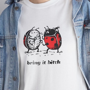 May include: White t-shirt featuring a graphic of two cartoon ladybugs with the text "bring it bitch" in black. The ladybugs have red and black shells. The shirt is worn with a blue denim jacket.