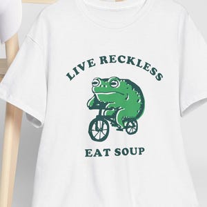 Puede incluir: Camiseta blanca con una rana verde montando una bicicleta y el texto "Live Reckless Eat Soup".