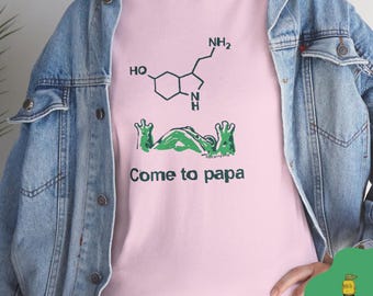 May include: Pale pink t-shirt featuring a green frog design and the words "Come to papa". A black and green chemical structure is above the frog. The t-shirt is made of a soft fabric.