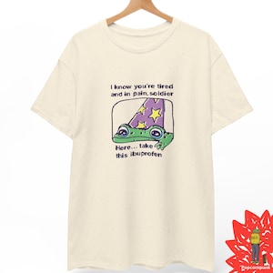 Può includere: Una t-shirt a maniche corte color crema con una stampa grafica di una rana verde che indossa un cappello da mago viola con stelle gialle. Il testo recita: I know you're tired and in pain, soldier. Here... take this ibuprofen. Il tessuto è maculato.