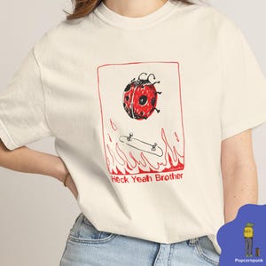 May include: Off-white t-shirt with a red and black ladybird above a skateboard, with flames and the text "Heck Yeah Brother" in red. The design has a playful, hand-drawn aesthetic.