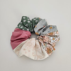 Op de afbeelding: Een veelkleurige stoffen scrunchie met een bloemenpatroon. De scrunchie is gemaakt van vijf verschillende stoffen, waaronder een groene stof met witte stippen, een lichtblauwe stof met een bloemenpatroon, een roze stof, een witte stof met borduurwerk en een perzikkleurige stof met een bloemenpatroon.